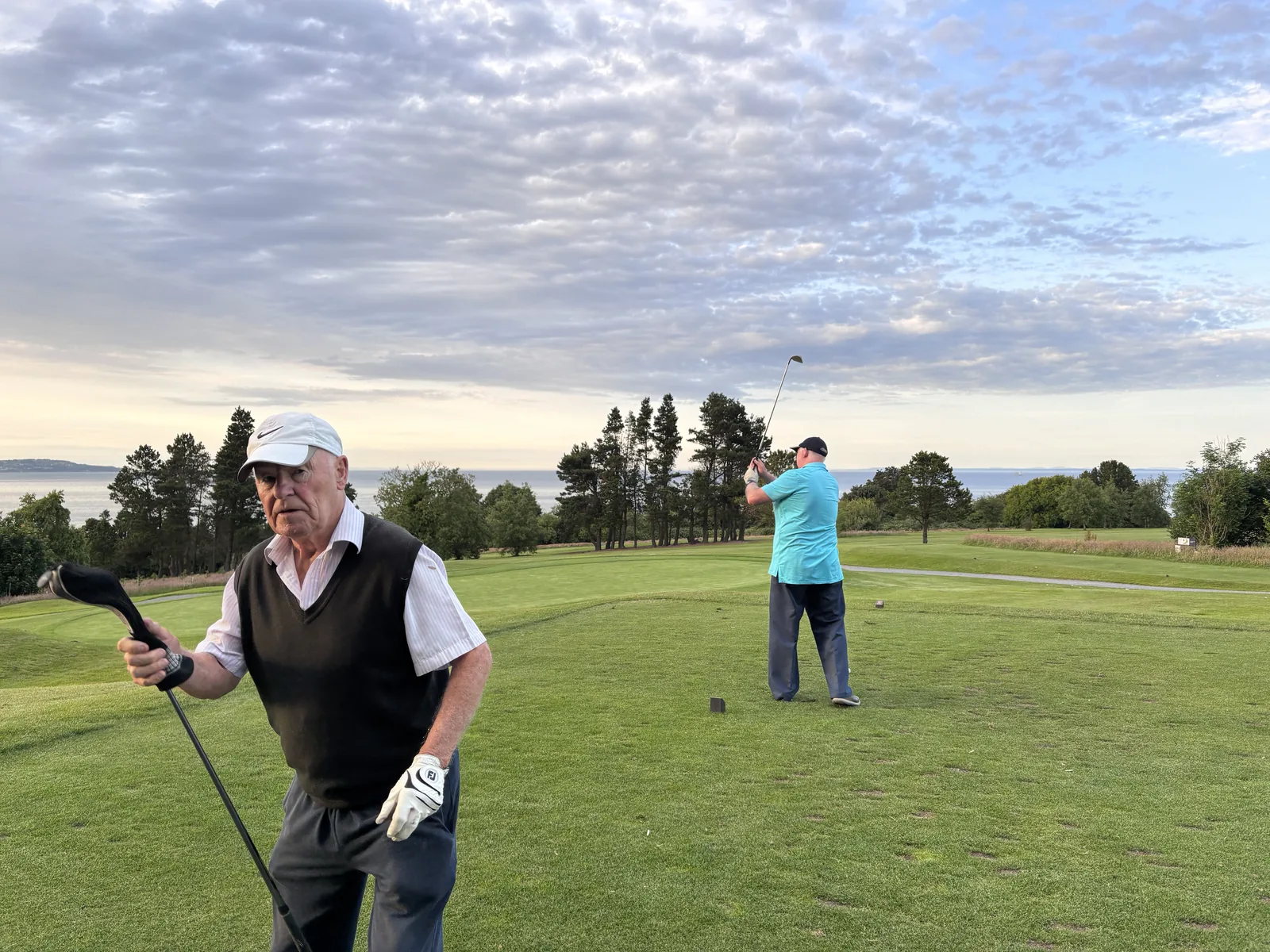 An older golfer in a flat cap walks off the tee holding his club while a younger playing partner finishes his swing behind him, on a coastal course at dusk.